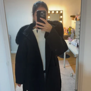 Svart kappa med fuskpäls och bälte - Snygg svart kappa med stor krage i fuskpäls och knytbälte i midjan. Jackan har en klassisk och stilren design med lång ärm och markerad axel. Perfekt för kyliga dagar när du vill känna dig extra uppklädd. Kappan är kashmir 
