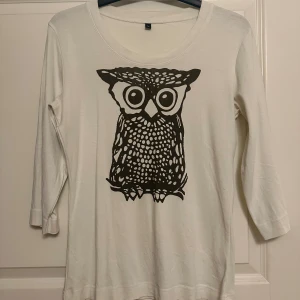 Vit långärmad tröja med uggletryck - Vit långärmad tröja med ett stort svart uggletryck på framsidan. Tröjan har rund halsringning och trekvartsärm, vilket ger en avslappnad och cool vibe. Perfekt för dig som gillar unika prints och vill sticka ut lite extra.