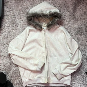 Vit/Grå Zip Hoodie med fuskpäls Storlek L - Vit ziphoodie med mjuk fuskpäls runt luvan. Jackan har dragkedja framtill, två fickor och broderad logga på bröstet. Materialet är bomull och polyester, perfekt för dig som gillar chill och streetwear-vibe. Tjock och varm perfekt för nutidens väder