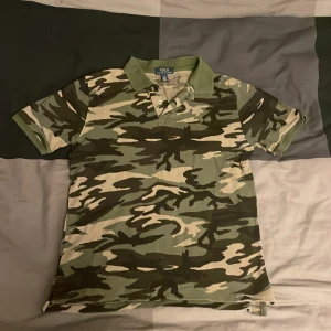 Camouflage pikétröja från Ralph Lauren - Säljer en grön och beige camouflage pikétröja från Polo Ralph Lauren. Klassisk krage, korta ärmar och broderad logga på bröstet. Tillverkad i mjuk bomull, perfekt för dig som gillar streetwear och vill sticka ut med en unik look. Skriv om intresse. Vi kan prata om priset.