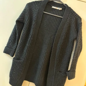 Svart stickad kofta från 157 - Mysig svart stickad kofta från 157 med grovstickad struktur, långa ärmar och två fickor framtill. Perfekt att slänga på sig när det är kyligt ute. Koftan har en öppen front och bred ribbad kant längs öppningen.