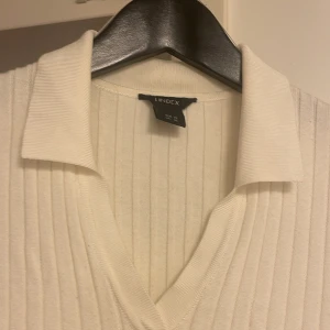 Vit ribbad pikétröja från Lindex - Vit ribbad pikétröja från Lindex med klassisk krage och v-ringning. Tröjan har trekvartsärm och är figurnära i modellen. Perfekt för dig som gillar en clean och stilren look. Materialet är mjukt och stretchigt, vilket gör den superbekväm. Tröjan troligen passar även dig som har storlek S.