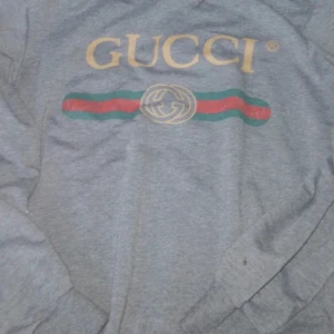 Grå långärmad Gucci tröja XL - Säljer en grå långärmad tröja från Gucci i storlek XL. Tröjan har det klassiska Gucci-logotyptrycket i guld framtill. 