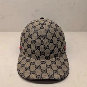 Gucci keps - Snygg keps från Gucci med klassiskt GG-mönster i svart på beige bakgrund. Kepsen har en böjd skärm och justerbar rem bak i svart skinn. På sidan finns en röd och marinblå rand som ger extra lyxig känsla. Perfekt för dig som vill sticka ut med designerstil.