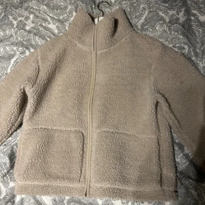 Beige teddyjacka från 157, storlek S - Mysig beige teddyjacka från 157 med hög krage och dragkedja framtill. Jackan har två stora fickor och är fodrad på insidan för extra komfort. Perfekt för kyliga dagar när du vill vara både varm och stilren.