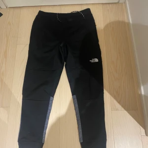 Svarta och grå täckbyxor från The North Face - Säljer ett par svarta och grå täckbyxor från The North Face med logga på benet. Byxorna har snörning i midjan och är tillverkade i en kombination av polyester och syntetmaterial. Perfekta för kalla dagar och outdoor-aktiviteter.