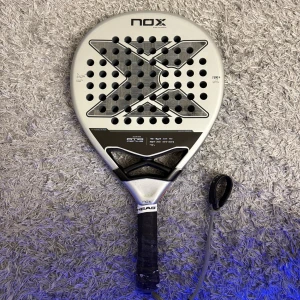NOX AT10 Genius 18K Padelracket - NOX AT10 Genius 18K padelracket i silver och svart. En spricka syns upptill på ramen, vilket kan påverka skick och spelbarhet minimalt och därmed låga prisintervallet. Dock går att reparera om man vill. 