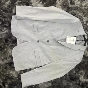 Ljusgrå kavaj Selected Homme - Snygg ljusgrå kavaj från Selected Homme med klassisk passform och två knappar framtill. Kavajen har diskreta fickor och stilren krage, perfekt för dig som gillar en clean och modern look. Materialet är mjukt och lätt, vilket gör den bekväm att bära.