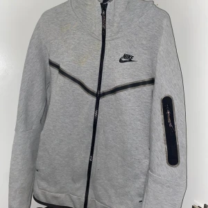 Grå Nike Tech - Snygg grå hoodie från Nike med svart logga på bröstet och hel dragkedja framtill. Tröjan har huva, svarta detaljer längs bröstet och en praktisk ficka med dragkedja på ärmen. Perfekt för chill eller när du är på språng.