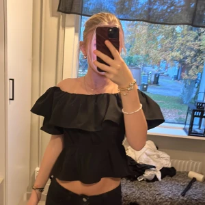Svart offshoulder volangtopp - Säljer en svart offshoulder topp med bred volang runt axlarna och lätt utsvängd nederdel. Toppen är croppad och har en feminin, trendig look som passar perfekt till jeans eller kjol. Materialet känns mjukt och följsamt.