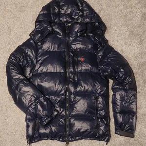 Mörkblå dunjacka Polo Ralph Lauren XS - Mörkblå dunjacka från Polo Ralph Lauren i storlek XS. Jackan har huva, dragkedja framtill och flera fickor med dragkedja. Klassisk röd Polo-logga broderad på bröstet. Perfekt för kalla dagar och riktigt snygg streetstil.