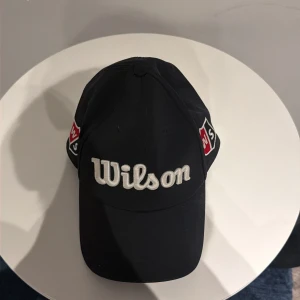 Svart Wilson keps med broderad logga - Snygg svart keps från Wilson med vit broderad logga framtill och bak. Kepsen har klassisk form, justerbar snapback och mesh på insidan för extra ventilation. Röd och vit Wilson-märke på sidorna ger en sportig vibe. Perfekt för en runda golf