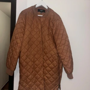 Quiltad brun jacka Vero Moda Curve - Quiltad lång jacka från Vero Moda Curve i en snygg brun färg. Jackan har rund hals, långa ärmar med muddar, tryckknappar framtill och dragkedja i sidan. Materialet är glansig polyester och jackan har en loose passform som ger en chill vibe.