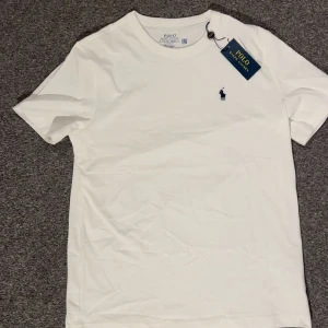 Vit t-shirt från Polo Ralph Lauren - Helt ny vit t-shirt från Polo Ralph Lauren med klassisk passform och korta ärmar. T-shirten har en rund hals och den ikoniska Polo-loggan broderad i marinblått på bröstet. Tillverkad i mjuk bomull för skön känsla och stilren look. Storlek S.