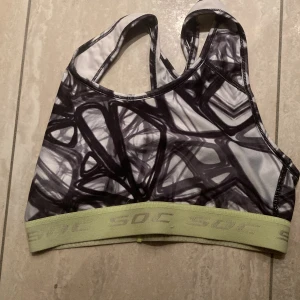 Svartvit sporttopp från SOC - Sporttopp från SOC i svartvitt abstrakt mönster med bred, ljusgrön resår nertill där det står 'SOC'. Modellen är ärmlös med brottarrygg och tillverkad i stretchigt material för bästa passform. Perfekt för träning och aktiva dagar.