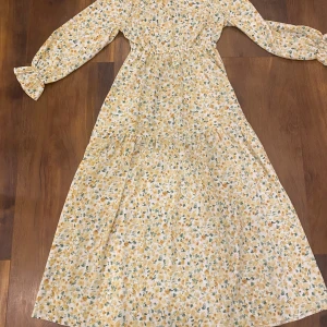 Lång blommig klänning med volangärm - Superfin långklänning med småblommigt mönster i gult och grönt på vit botten. Klänningen har långa ärmar med volangdetalj vid ärmslut och markerad midja för en feminin siluett. Perfekt för vår och sommar!