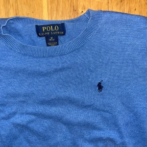 Blå stickad tröja från Polo Ralph Lauren - Snygg blå stickad tröja från Polo Ralph Lauren med klassisk rund hals och broderad logga på bröstet. Tröjan har ribbade muddar vid ärmslut och nederkant samt långärmad passform. Perfekt för dig som gillar stilrena och tidlösa plagg.