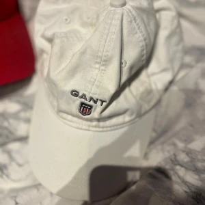 Vit keps från GANT - Snygg vit keps från GANT med broderad logga och sköld framtill. Klassisk böjd skärm och justerbar passform. Tillverkad i mjuk bomull som är skön att bära hela dagen. Perfekt för dig som vill ha en clean och stilren look.