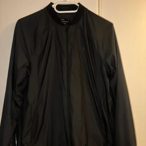 Svart vindjacka från Peak Performance - Svart vindjacka från Peak Performance i stilren design. Jackan har dragkedja framtill, diskret logga på bröstet och ribbad krage. Tillverkad i lätt och smidigt syntetmaterial, perfekt för blåsiga dagar. Passar dig som gillar sportig och clean look.