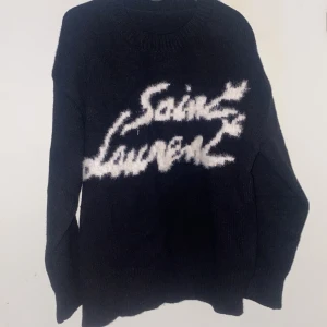 Svart stickad Saint Laurent tröja L - Säljer en svart stickad tröja från Saint Laurent med vit, fluffig logga framtill. Tröjan har rund hals och långa ärmar, perfekt för dig som gillar statement-plagg och exklusiv streetstyle.
