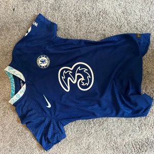 Chelsea FC blå Nike fotbollströja - Chelsea FC matchtröja i blått från Nike med klubbmärke på bröstet och stor vit sponsorlogga. Tröjan har korta ärmar, Dri-FIT ADV-material och en krage med ljusblå detaljer och grafiskt mönster. Perfekt för dig som vill representera Londonklubben.