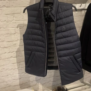 Svart dunväst från Moose Knuckles - Snygg svart dunväst från Moose Knuckles med diskret logga på bröstet. Västen har quiltad design, hög krage och två snedställda fickor framtill. Perfekt för lager-på-lager och ger en clean och modern look.