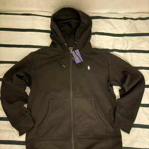 Svart hoodie från Polo Ralph Lauren - Svart hoodie från Polo Ralph Lauren med dragkedja, huva med snörning och klassisk broderad logga på bröstet. Tröjan har känguruficka framtill och är tillverkad i mjuk bomull. Perfekt för en chill och stilren look.