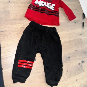 Svarta mjukisbyxor med röda ränder mickey mouse Disney  - Svarta mjukisbyxor med elastisk midja och ribbade muddar vid bensluten. På ena benet finns tre glansiga röda ränder som ger en sportig vibe. Perfekta för chill eller häng med kompisar. Materialet känns mjukt och bekvämt mot huden.Ett sött set bestående av en röd tröja med texten "Hello Mouse" och ett par svarta byxor med röda ränder på benen.