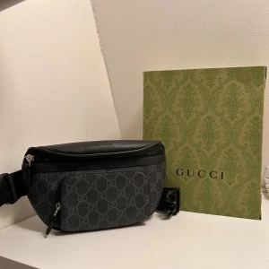 Svart magväska med GG-mönster - Snygg svart magväska med klassiskt GG-mönster i grått. Väskan har justerbart band, dragkedja och detaljer i skinn. Perfekt för att bära det viktigaste och ge din outfit en lyxig touch.