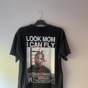 Svart Cactus Jack t-shirt Travis Scott - Svart t-shirt från Cactus Jack med stor grafisk print på ryggen och mindre tryck på bröstet. Motiv med texten 'LOOK MOM I CAN FLY' och Travis Scott-inspirerad design. Rund hals och korta ärmar, tillverkad i mjuk bomull.