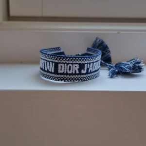 Blått Christian Dior J'Adior armband - Snyggt blått och vitt armband från Christian Dior med texten 'J'Adior'. Armbandet är vävt i tyg och har dekorativa tofsar i ändarna med knytning för justerbar passform (passar alla). Perfekt accessoar för att lyfta din outfit. Hör av er vid minsta fråga 🤍🙌