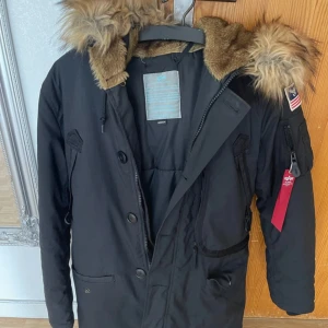 Svart parkajacka Alpha Industries M - Parkajacka från Alpha Industries i svart med stor huva och snygg fuskpäls. Jackan har flera fickor, patch med amerikansk flagga på ärmen och både dragkedja och knappar framtill. Perfekt för kalla dagar och har en cool streetstil. Använt 1 gång 