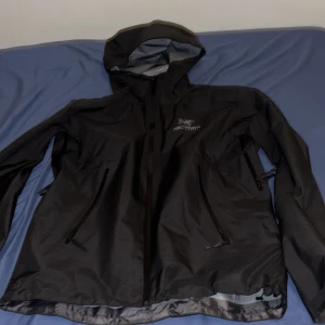 Svart Arc'teryx vindjacka med huva - Arc'teryx vindjacka i svart med stilren design och huva. Jackan har flera dragkedjeförsedda fickor och logga broderad på bröstet. Materialet är lätt och syntetiskt, perfekt för blåsiga dagar. Passformen är normal och jackan är långärmad.