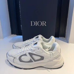 Vita Dior B30 sneakers med grå detaljer - Säljer ett par Dior B30 sneakers i vitt med gråa detaljer och CD-logga på sidan och hälen. Skorna har en chunky sula i svart och vitt, snörning och meshpaneler för extra komfort. Perfekta för dig som vill ha en lyxig och modern sneaker med streetkänsla.