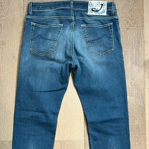 Blå jeans från Jacob Cohen - Snygga blå jeans från Jacob Cohen med klassisk femficksdesign och kontrastsömmar. Jeansen har en unik patch med blått mönster och J-logga bak i midjan. Materialet är mjukt denim och passformen är rak med normal midja. Perfekta för en avslappnad och stilren look.