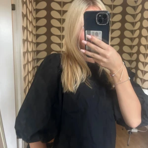 Svart blus med puffärm och rosetter - Supersöt svart blus med stora puffärmar och två vita rosetter på ryggen. Blusen har en boxig passform och är tillverkad i ett lätt bomullstyg. Perfekt för dig som vill sticka ut med en trendig och lekfull look.