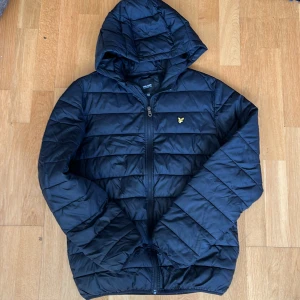Mörkblå pufferjacka från Lyle & Scott - Snygg mörkblå pufferjacka från Lyle & Scott med huva och gul broderad logga på bröstet. Jackan har quiltad design, dragkedja framtill och långa ärmar. Perfekt för kyliga dagar och har en clean, sportig vibe. Det finns en liten reva på ena ärmen och den är sliten vid muddarna, se bilder. Men det är inget som syns på håll eller stör funktionen. 