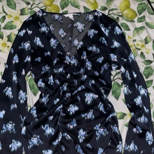 Blommig svart tröja från H&M - Svart tröja från H&M med blått och lila blommönster, v-ringad och med långa ärmar. Snyggt rynkad framtill och tillverkad i glansigt, mjukt material som känns lyxigt. Perfekt för dig som gillar en feminin och trendig look.