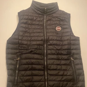 Svart dunväst från Colmar - Snygg svart dunväst från Colmar med quiltad design och hög krage. Västen har dragkedja framtill, två fickor med dragkedja och Colmar-logga på bröstet. Perfekt för lager-på-lager och streetstyle.