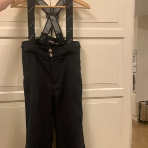 Svarta täckbyxor från Tuxer m hängslen, 134/140 - Svarta täckbyxor från Tuxer med justerbara hängslen och justerbar midja. Dragkedjor vid fickorna. Byxorna har reflexdetalj på benet och är fodrade för extra värme. Några små defekter på fållen längst ner och på vänster knä, därav priset, inget som stör helhetsintrycket. 