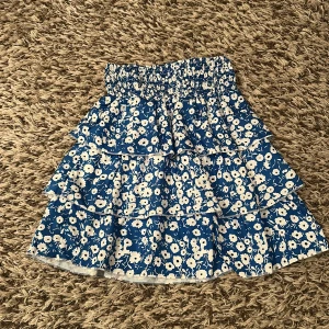 Blommig blå volangkjol med resår - Supersöt kort kjol med blå botten och vita blommor. Kjolen har flera volanger och ett brett resårband i midjan som gör den bekväm och enkel att bära. Materialet känns lätt och luftigt, perfekt för sommaren💗