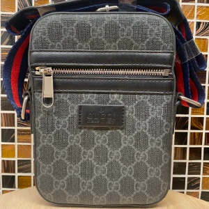 GG axelväska med GG-mönster - Snygg axelväska GG i mörkgrått med klassiskt GG-mönster över hela väskan. Väskan har en dragkedja framtill och justerbar axelrem i blått och rött. Tillverkad i canvas med detaljer i svart skinn. Perfekt för dig som vill ha en stilren och lyxig look.