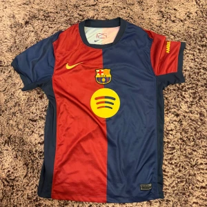 FC Barcelona matchtröja Nike M - 🚨MAN KAN BARA TA BORT NAMNET HEMMA🚨Säljer en FC Barcelona matchtröja från Nike i storlek M. Tröjan har klassiska rödblå vertikala fält, gult tryck med nummer 10 och namnet Anton på ryggen, samt Spotify och UNHCR-loggor. Tillverkad i lätt Dri-Fit material som andas, perfekt för fotboll eller träning.