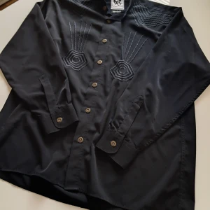 Svart skjorta med broderi från Antonio Archini - Svart skjorta från Antonio Archini med unika geometriska broderier på bröstet och axlarna. Skjortan har klassisk krage, knappar framtill och långa ärmar med manschett. Materialet känns som tjock bomull och polyester, kan vara tom siden, eftersom handtvätt rekommenderas. Normal Passformen. Perfekt för dig som gillar detaljer och stil. 