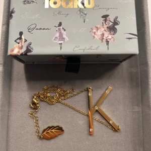 Guldfärgat halsband med bokstav K - Stilrent halsband från i iaoku guldfärg med en stor bokstav K dekorerad med små glittrande stenar. Kedjan har även ett snyggt lövformat hänge som extra detalj. Perfekt accessoar för dig som vill sticka ut med din initial.