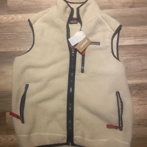 Beige fleeceväst från Patagonia - Beige fleeceväst från Patagonia med marinblå detaljer och röda dragkedjor. Västen har hög krage, två fickor med dragkedja och en bröstficka. Perfekt för lager-på-lager och outdoor-vibe. Klassisk logga på bröstet och mjukt, mysigt material.
