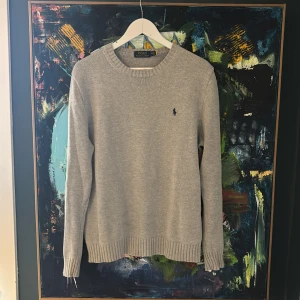 Grå stickad tröja från Polo Ralph Lauren - Snygg grå stickad tröja från Polo Ralph Lauren med klassisk rund hals och diskret broderad logga på bröstet. Tröjan har ribbade muddar vid ärmslut och nederkant, perfekt för dig som gillar stilrena och tidlösa plagg.