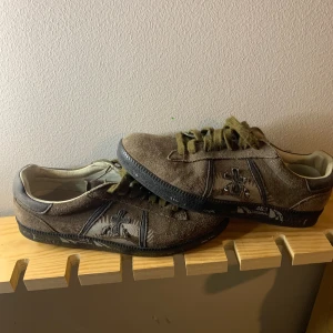 Premiata skor - Snygga bruna sneakers från Premiata i mjuk mocka med broderad logga på sidan och svarta detaljer. Skorna har snörning, svart platt sula och rund tå. Perfekta för dig som gillar en stilren men unik look. Storlek 40 men passar större. Skulle säga 41