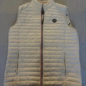 Vit dunväst från JOTT - Snygg vit dunväst från JOTT med horisontella quiltade sömmar och coolt tryck med scooter på bröstet och ryggen. Västen har dragkedja med röd och blå detalj, två fickor med dragkedja och stårkrage. Perfekt för lager-på-lager under kyliga dagar.