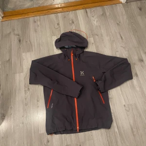 Mörkgrå Haglöfs vindjacka S - Snygg mörkgrå vindjacka från Haglöfs i storlek S. Jackan har huva, orange dragkedjor och detaljer, samt flera praktiska fickor. Tillverkad i slitstark Gore-Tex polyester, perfekt för utomhusaktiviteter. Stilren design med Haglöfs-logga på bröstet.